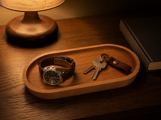 Cherry wood catch all tray on a walnut nightstand holding a leather-strap watch and keys on a leather fob, lit by a warm bedside lamp