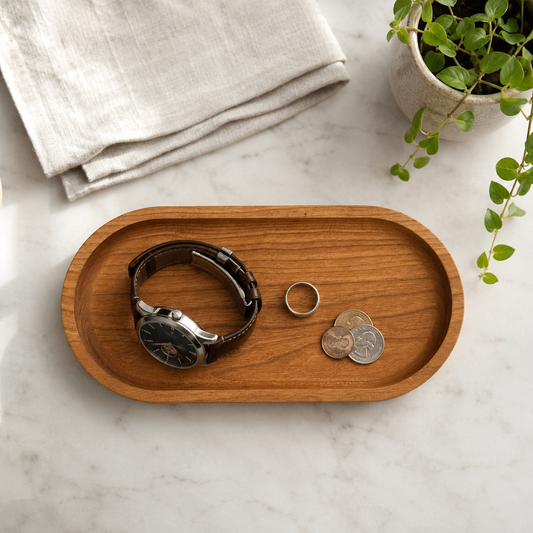 Overhead view of the cherry catch all tray on a marble dresser top holding a watch, a ring, and loose coins beside folded linen and a trailing plant