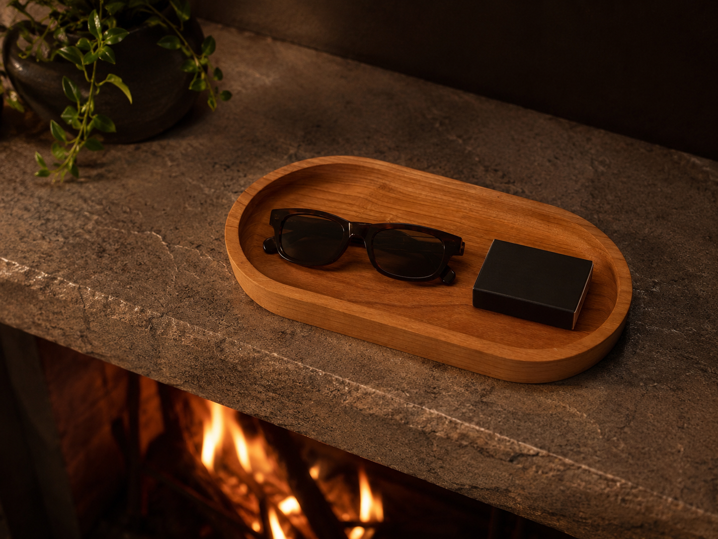 Cherry catch all tray on a stone fireplace mantle holding folded sunglasses and a matchbox, with warm firelight glowing below